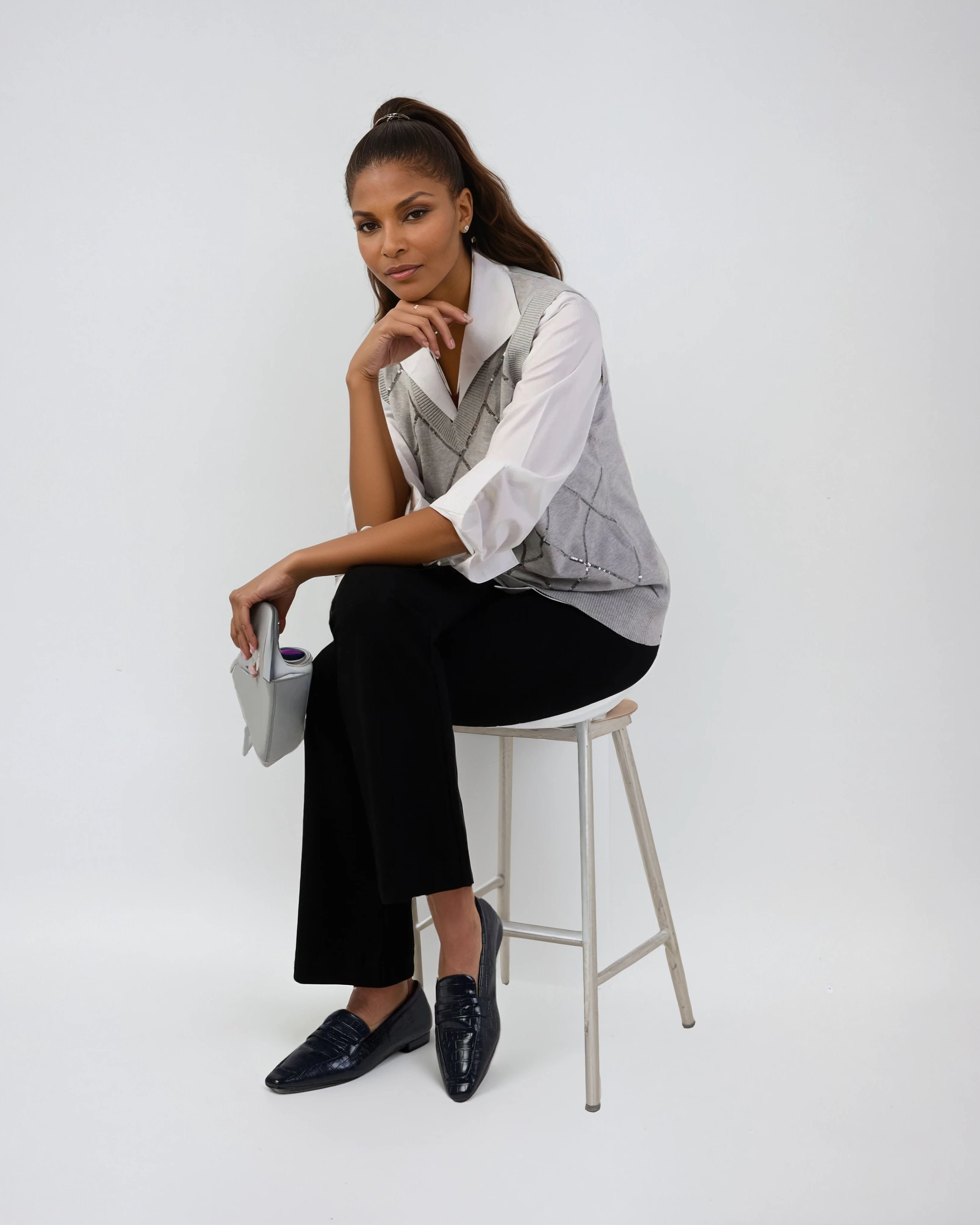 Woman sitting on a stool wearing a white shirt, gray vest, black pants, and black shoes against a white background.