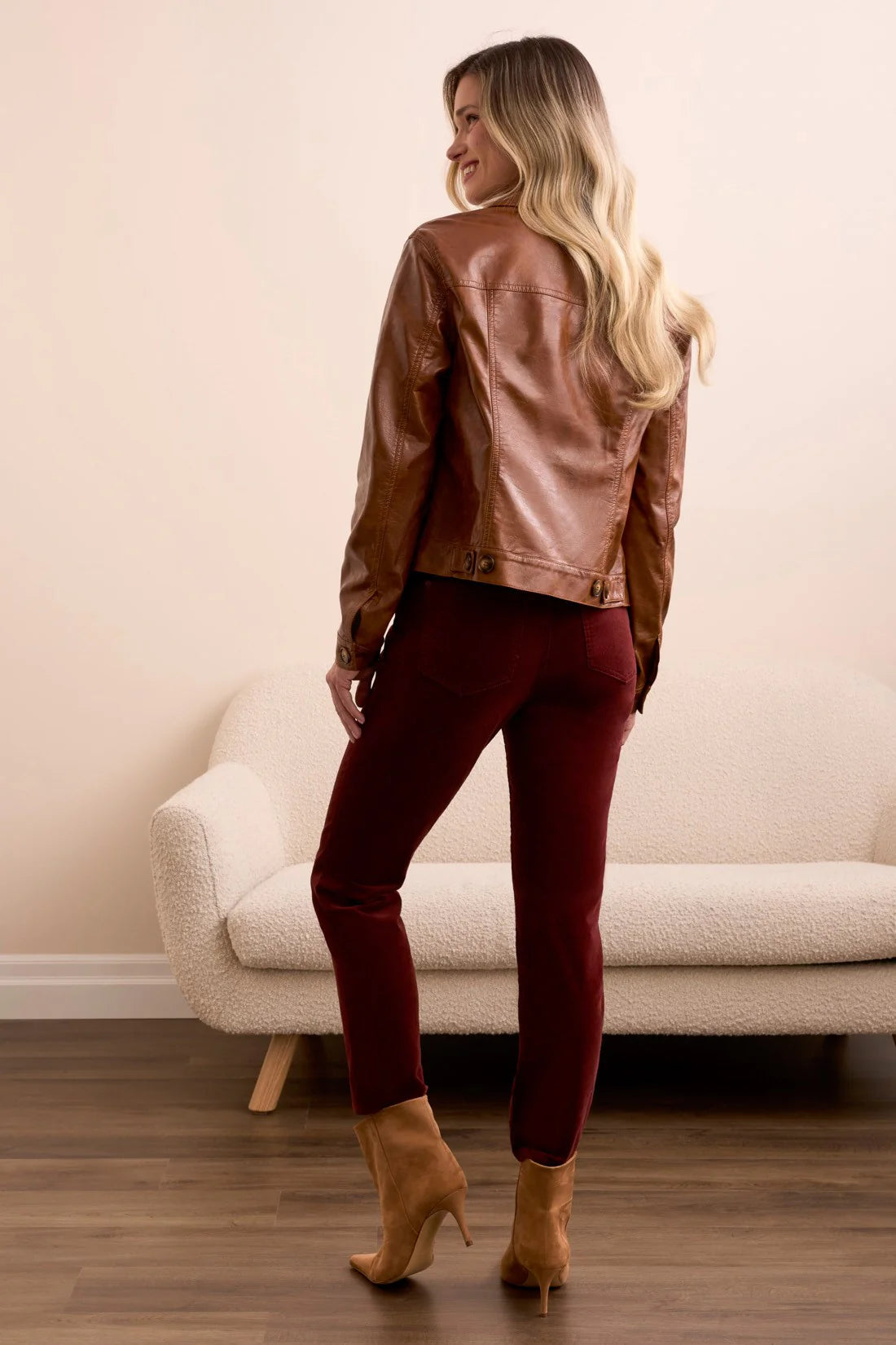 Woman wearing a brown leather jacket and red pants standing in a room with a beige sofa.