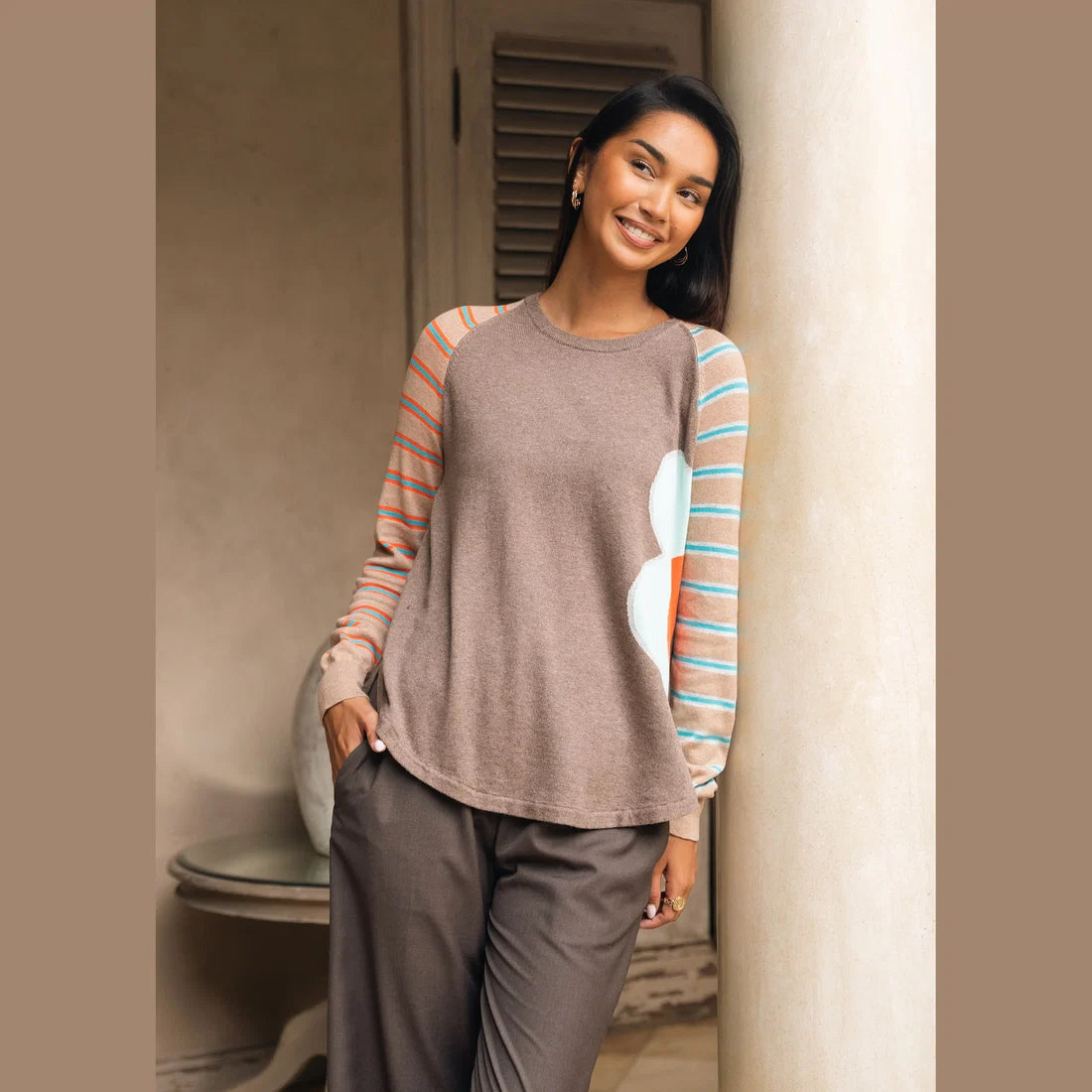 A woman modeling a long-sleeve crewneck top with a solid brown body, peach and turquoise striped sleeves, and a large light blue and coral abstract flower on the right side Zaket & Plover Walnut Bright Side Sweater