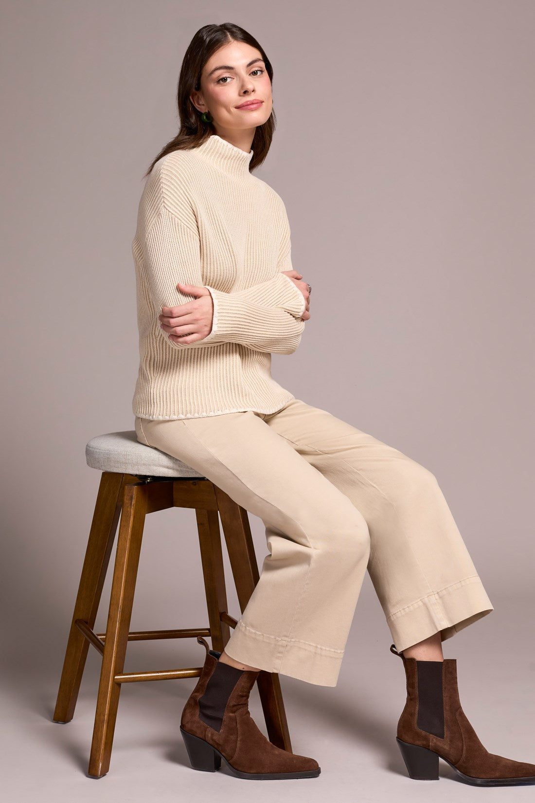 Woman sitting on a stool wearing a beige sweater, pants, and brown boots against a plain background
