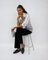 Woman sitting on a stool wearing a white shirt, gray vest, black pants, and black shoes against a white background.