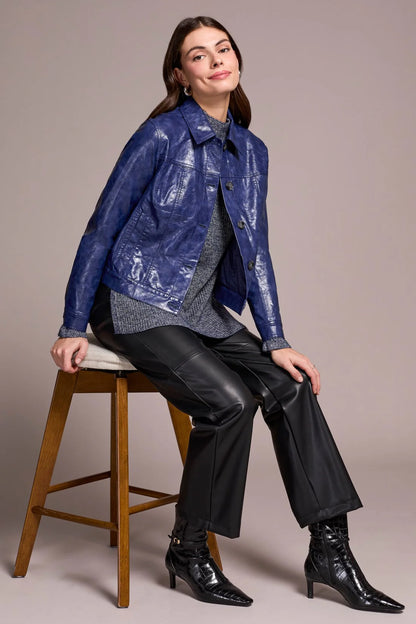 Woman sitting on a stool wearing a blue metallic jacket and black pants against a neutral background