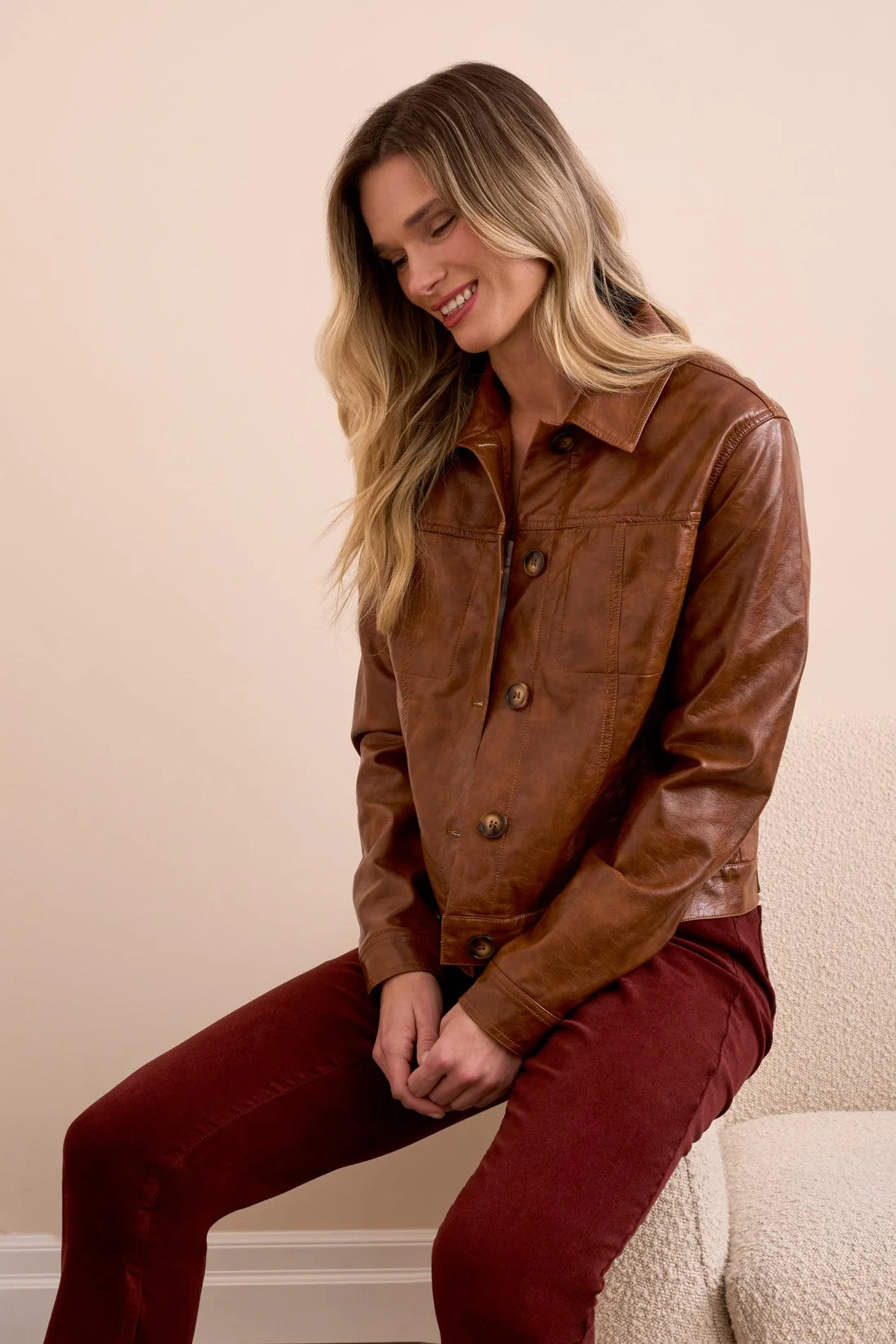 Woman wearing a brown leather jacket and burgundy pants sitting on a beige couch.