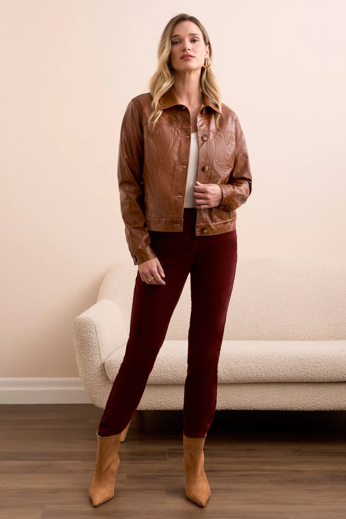 Woman wearing a brown jacket, white shirt, red pants, and tan boots standing in a room with a beige wall and white chair.