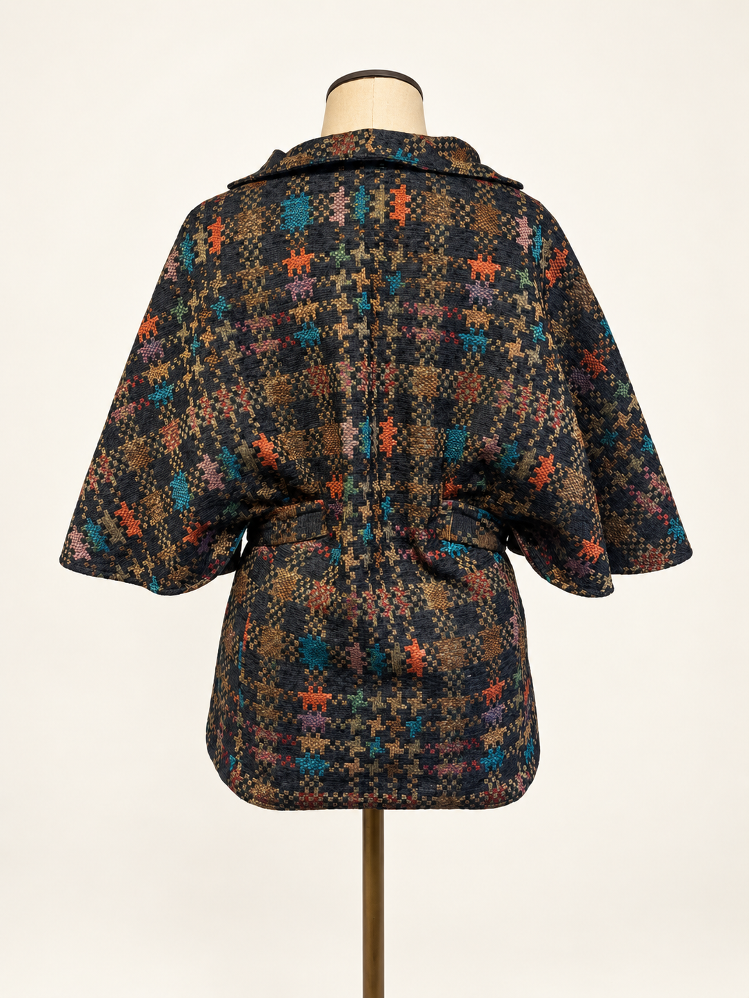 A short hooded cape-style jacket with a tie waist belt, displayed on a dress form. The fabric features a dense multicolor plaid and houndstooth pattern with black, brown, orange, and teal tones.
