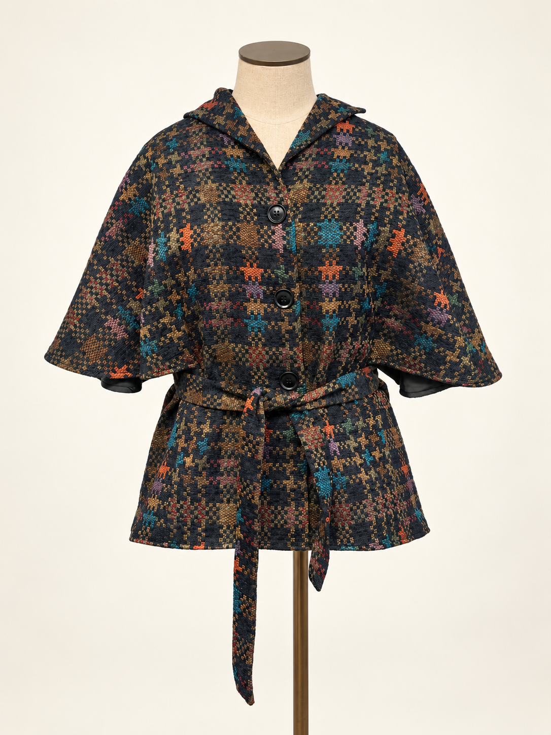 A short hooded cape-style jacket with a tie waist belt, displayed on a dress form. The fabric features a dense multicolor plaid and houndstooth pattern with black, brown, orange, and teal tones.