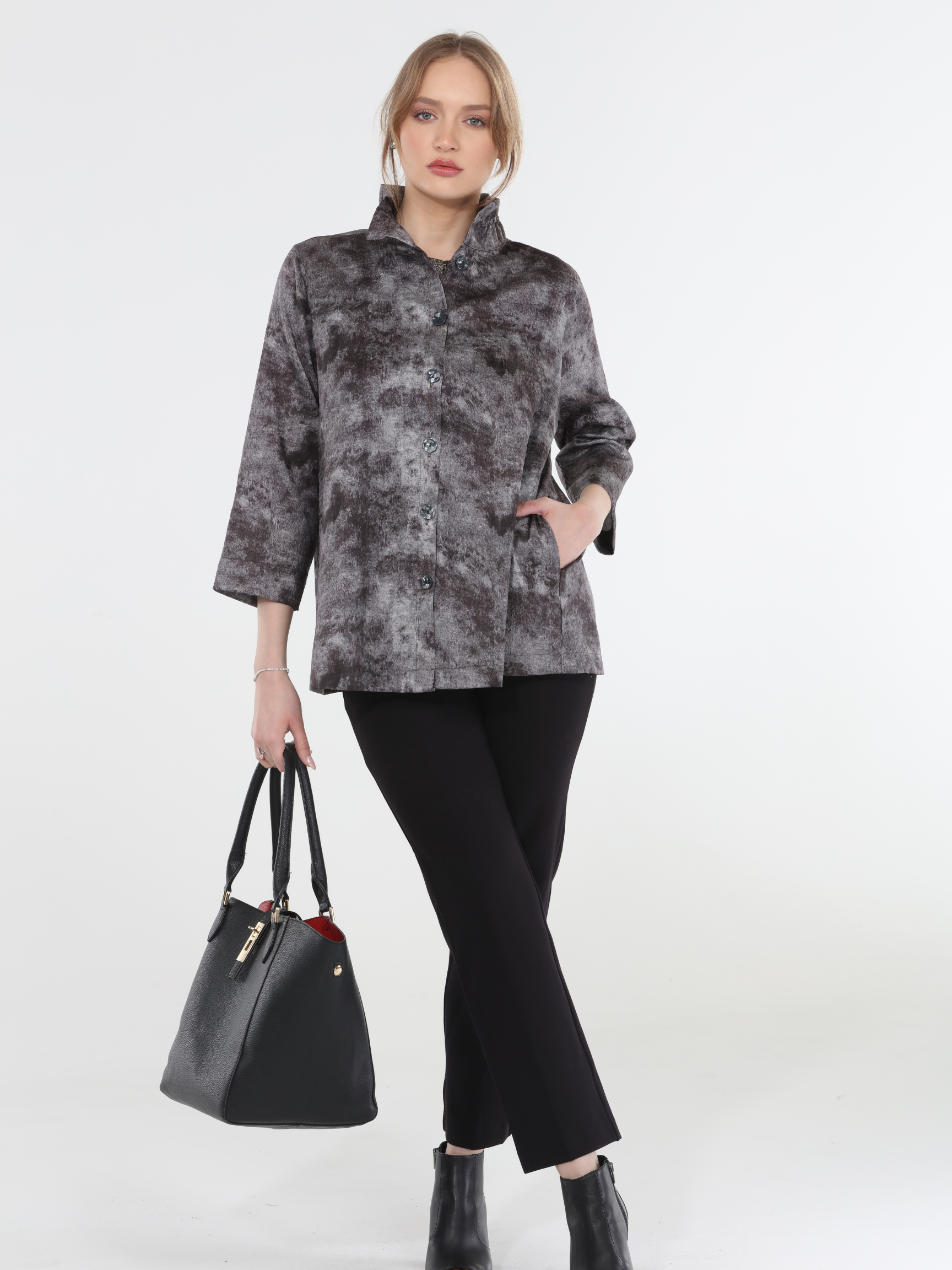 Woman wearing a gray patterned jacket and black pants, holding a black handbag on a white background