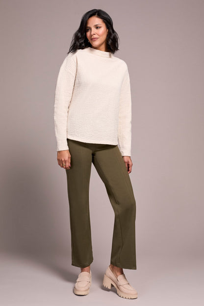 Woman wearing a cream sweater and green pants against a plain background