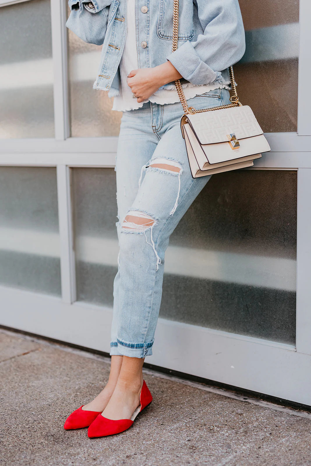 Person wearing a light blue denim jacket, ripped jeans, and red flats, holding a beige handbag against a glass wall.