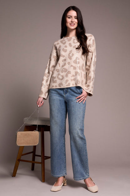 Woman wearing a patterned sweater and jeans standing next to a wooden stool with a beige bag.