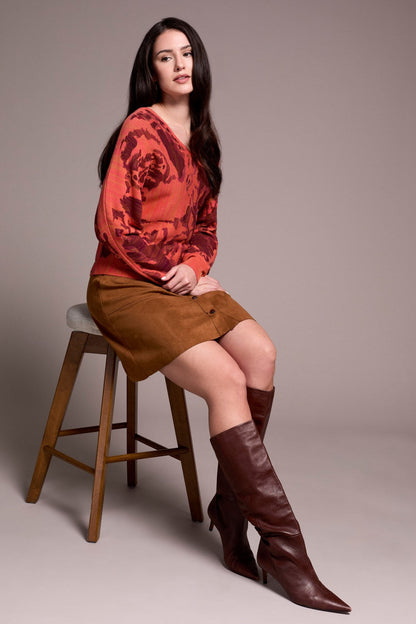 Woman sitting on a stool wearing an orange top and brown skirt with brown knee-high boots against a plain background