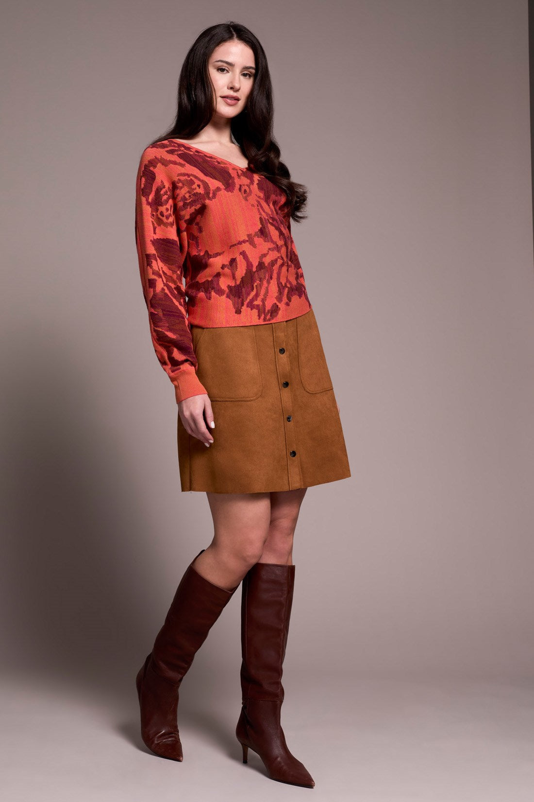 Woman wearing a red patterned sweater and brown skirt with knee-high boots on a plain background