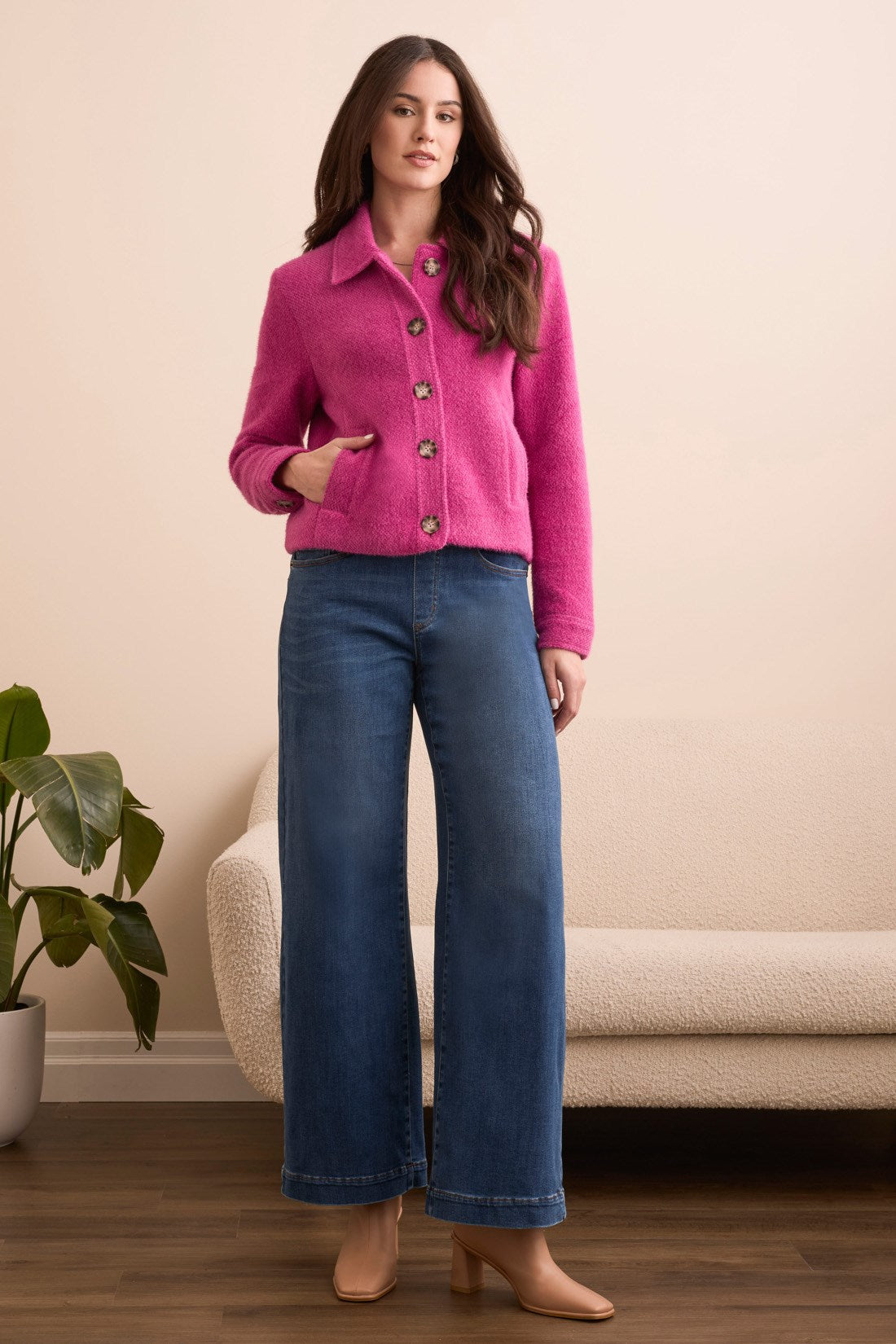 Woman wearing a pink jacket and blue jeans standing in a room with a plant and sofa.