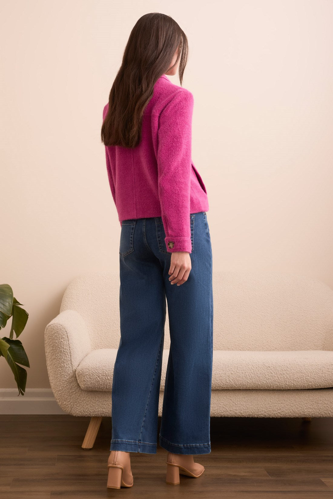 Woman wearing a pink sweater and blue jeans standing in a room with a beige couch and plant.