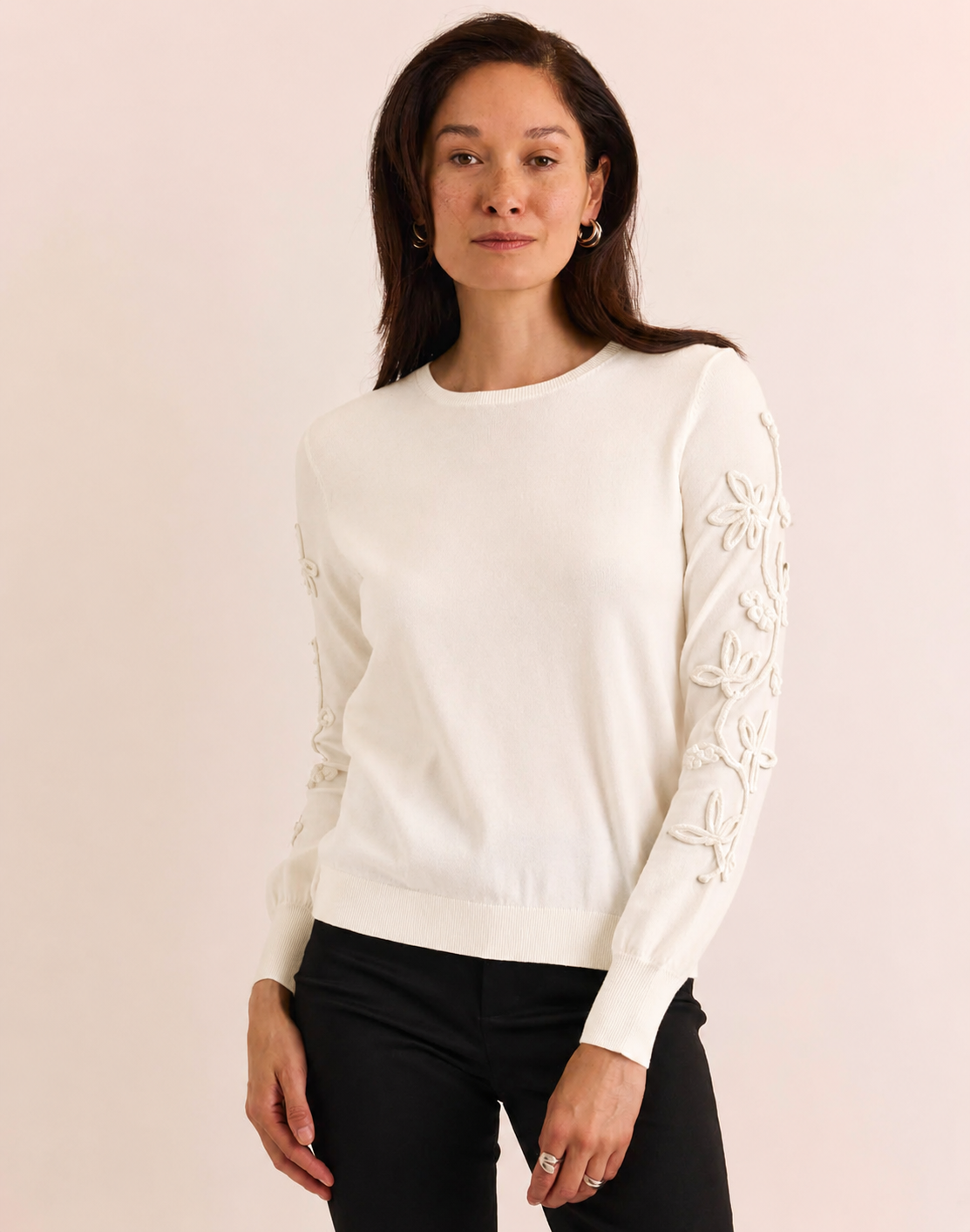 A woman modeling a cream-colored crewneck sweater with intricate floral embroidery on the sleeves