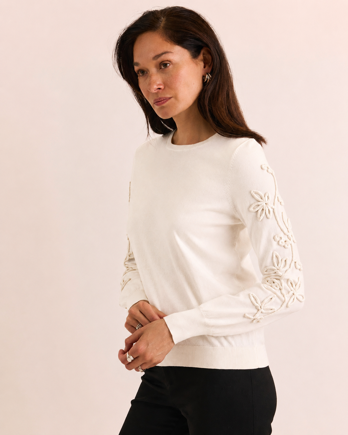 A woman modeling a cream-colored crewneck sweater with intricate floral embroidery on the sleeves